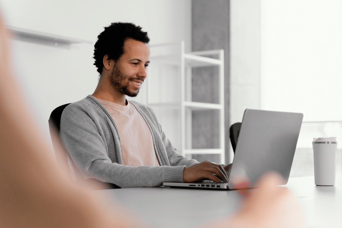 Homem jovem sentado em cadeira, sorrindo e digitando em notebook em ambiente de escritório moderno e bem iluminado