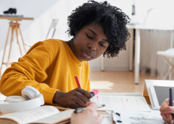 Jovem com camiseta amarela escrevendo em caderno ao lado de fones e tablet em mesa