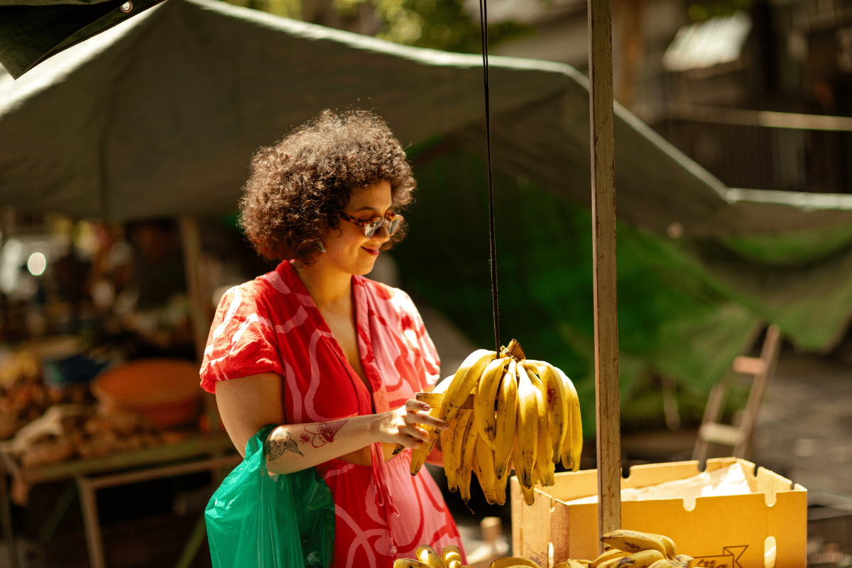 Pessoa com vestido vermelho e óculos segurando bananas penduradas em feira ao ar livre