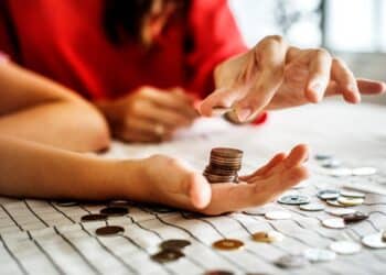 Mãos de uma pessoa colocando moedas de metal em uma pilha. Outras moedas estão espalhadas sobre uma mesa, simbolizando economia e colecionismo.