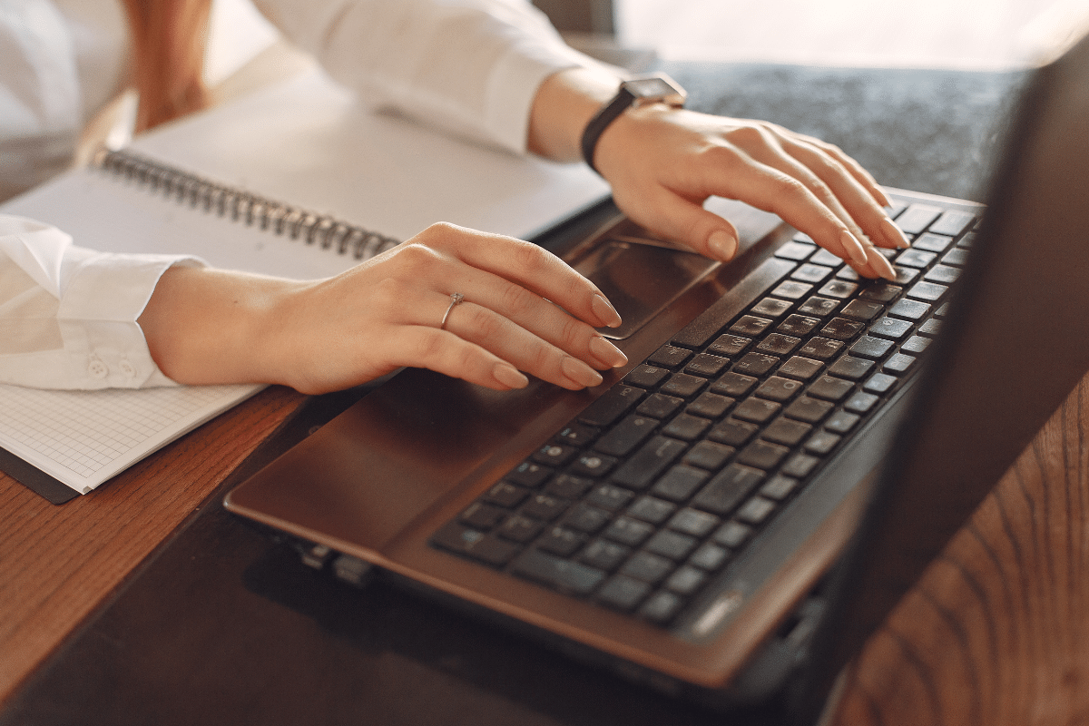 Duas mãos femininas digitando em teclado de notebook sobre mesa com caderno aberto ao lado