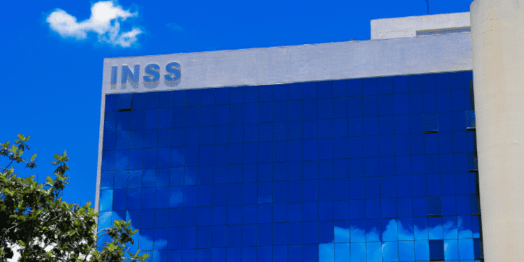Imagem de um prédio do INSS com fachada de vidro azul refletindo o céu e nuvens.