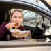 Mulher comendo macarrão dentro do carro com janela aberta.