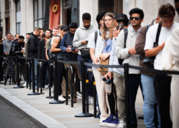 Diversas pessoas de diferentes idades e estilos esperando em fila organizada em uma calçada urbana