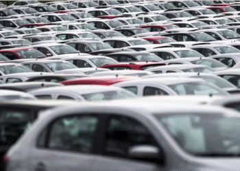 Estacionamento cheio com carros novos brancos, cinzas e vermelhos alinhados lado a lado em grande quantidade.