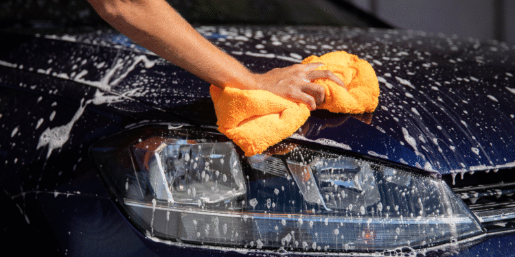 Pessoa lavando carro azul escuro com pano amarelo e sabão, foco no farol e espuma