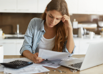 Mulher sentada em mesa com expressão preocupada verificando documentos financeiros ao lado de laptop e calculadora