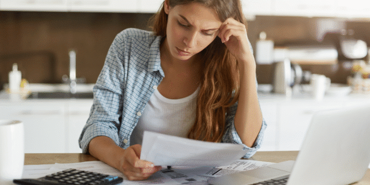 Mulher sentada em mesa com expressão preocupada verificando documentos financeiros ao lado de laptop e calculadora