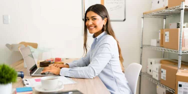 Mulher sorridente trabalhando no computador em um escritório, com mesa organizada e ambiente de trabalho moderno.