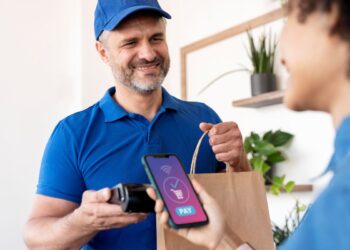 Entregador sorridente com uma maquininha de cartão, enquanto um cliente aproxima o smartphone para efetuar um pagamento por aproximação, representando a conveniência dos aplicativos de delivery.