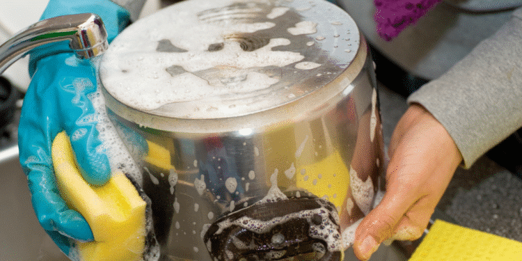 Pessoa lavando panela de alumínio com esponja amarela e luva azul na pia