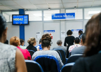 Pessoas sentadas aguardando atendimento em sala de perícia médica com painel eletrônico de senhas.