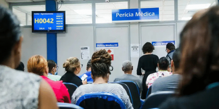 Pessoas sentadas aguardando atendimento em sala de perícia médica com painel eletrônico de senhas.