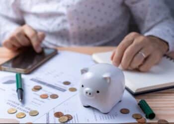 Pessoa fazendo cálculos com smartphone em uma mesa com papéis, dinheiro e um pequeno cofre de porquinho, representando o planejamento financeiro para economizar.