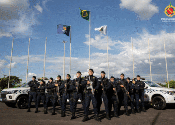 Grupo de policiais militares do Distrito Federal posando em formação com armas e viaturas ao fundo