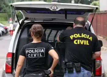 Dois policiais civis de costas com uniformes pretos próximos a viatura em operação policial