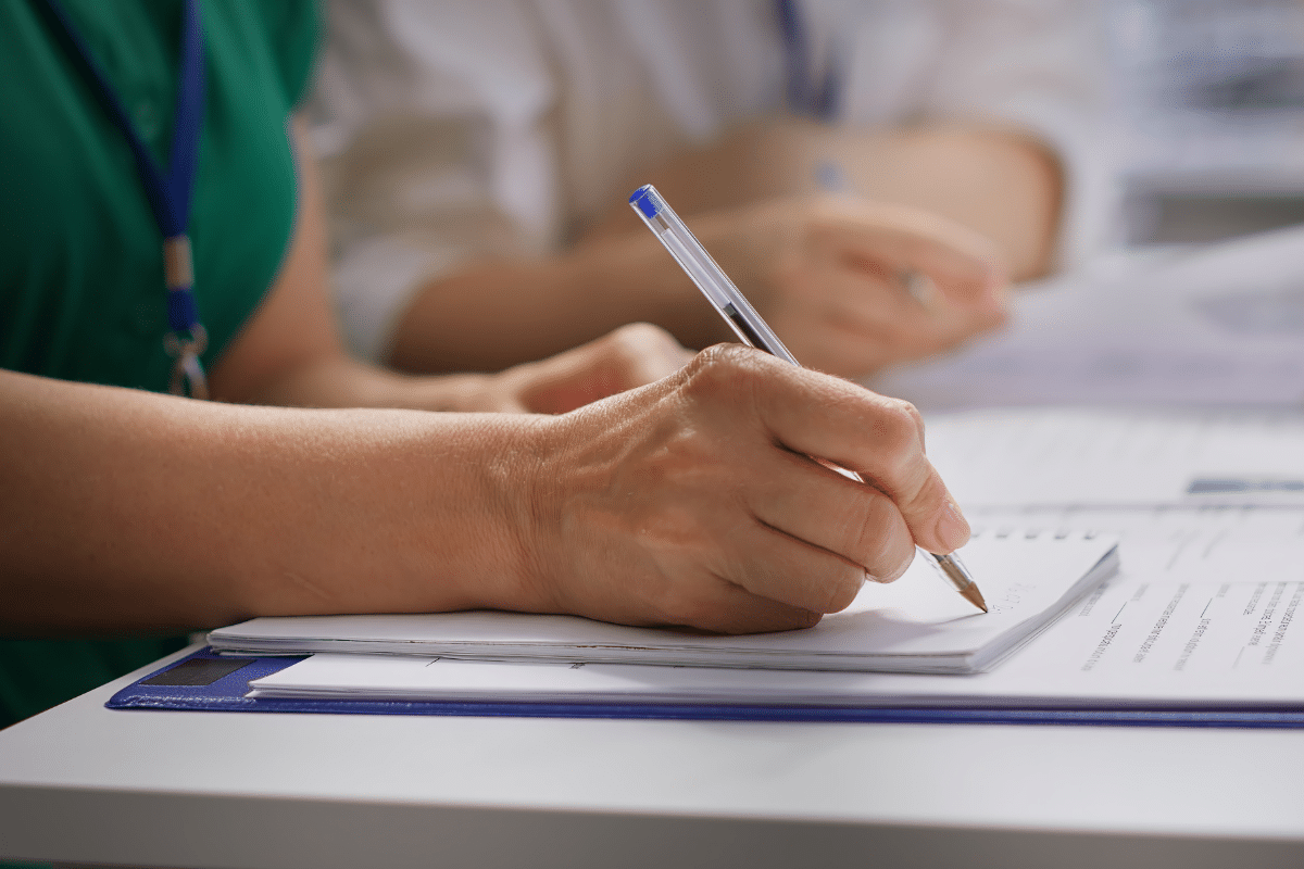 Pessoa escrevendo em bloco de notas com caneta azul sobre mesa branca, foco na mão
