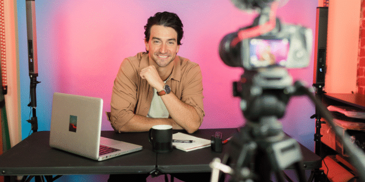 Homem sorridente gravando vídeo em estúdio com câmera, notebook e luzes coloridas ao fundo