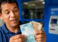 Homem conta notas de real próximo a um caixa eletrônico, em referência à projeção do salário mínimo de R$ 1.741 para 2027.