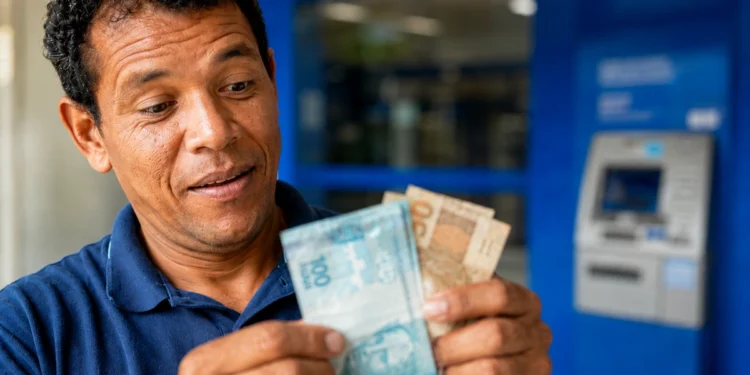 Homem conta notas de real próximo a um caixa eletrônico, em referência à projeção do salário mínimo de R$ 1.741 para 2027.