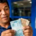 Homem conta notas de real próximo a um caixa eletrônico, em referência à projeção do salário mínimo de R$ 1.741 para 2027.