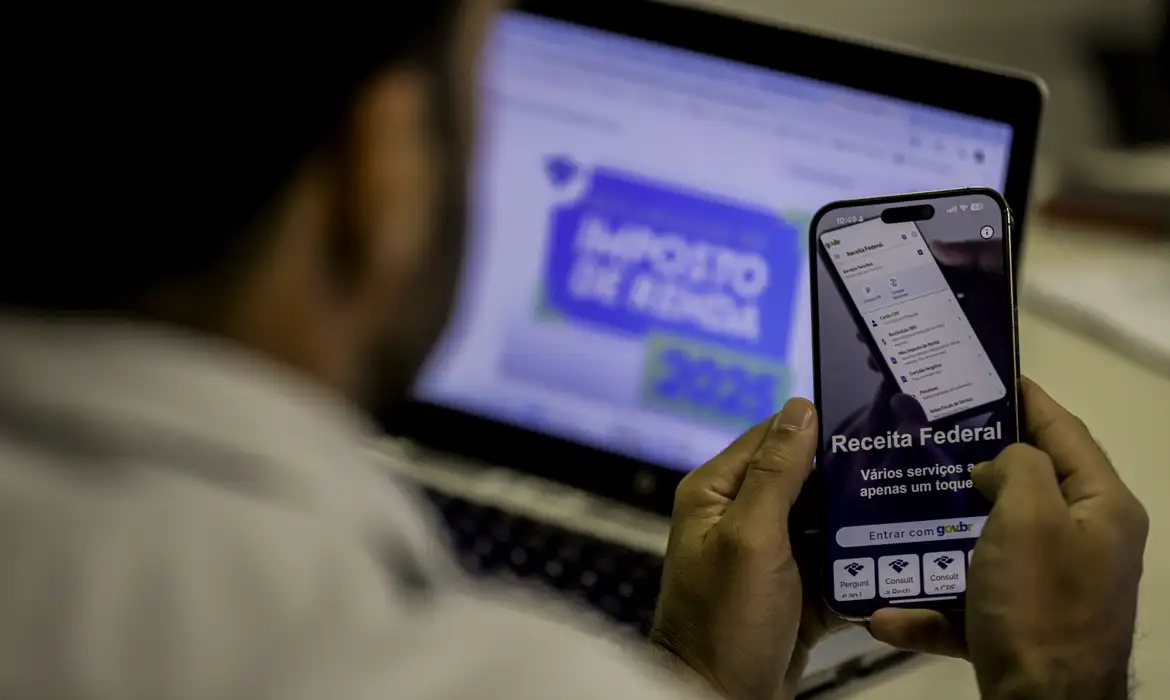 Pessoa utilizando o aplicativo da Receita Federal no celular enquanto consulta informações sobre o Imposto de Renda 2025 no computador.