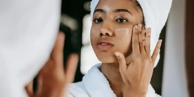 Mulher com toalha na cabeça aplicando creme facial na pele após o banho, em uma rotina de cuidados pessoais.