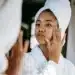 Mulher com toalha na cabeça aplicando creme facial na pele após o banho, em uma rotina de cuidados pessoais.