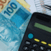 A imagem mostra várias cédulas de real ao lado de uma calculadora preta e uma caneta prata, colocadas sobre uma planilha com números e valores. A cena representa o cálculo financeiro, gerenciamento e planejamento econômico.