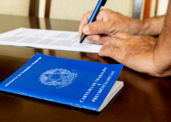 Mãos de pessoa preenchendo documento ao lado de carteira de trabalho azul sobre mesa de madeira