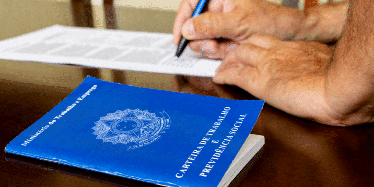 Mãos de pessoa preenchendo documento ao lado de carteira de trabalho azul sobre mesa de madeira