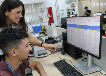 Duas pessoas em ambiente de escritório interagem com planilha aberta em tela de computador.