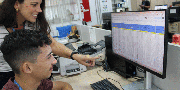 Duas pessoas em ambiente de escritório interagem com planilha aberta em tela de computador.