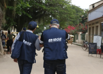 Dois agentes do IBGE com coletes azuis caminham em rua arborizada, coletando dados