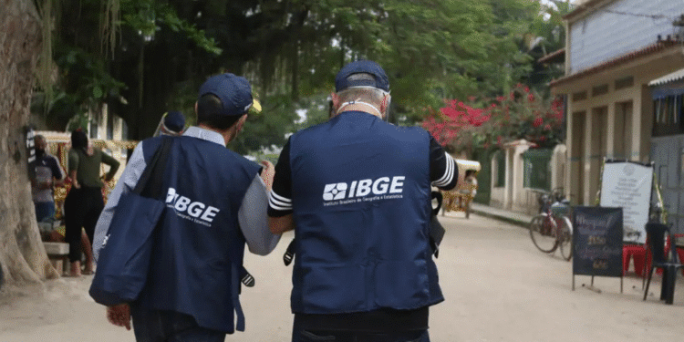 Dois agentes do IBGE com coletes azuis caminham em rua arborizada, coletando dados