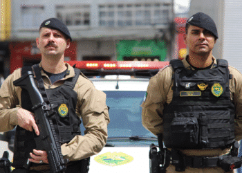 Dois policiais militares do Paraná com uniformes táticos e capacetes, um segurando fuzil em posição de frente a viatura policial