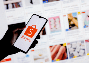 Mão segurando celular com logo da Shopee na frente de tela com diversas imagens de produtos à venda