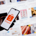Mão segurando celular com logo da Shopee na frente de tela com diversas imagens de produtos à venda