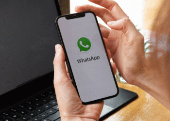 Pessoa segurando celular com tela mostrando o aplicativo WhatsApp aberto, ao lado de notebook