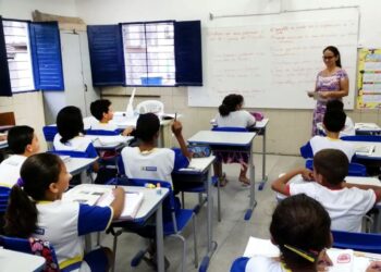 Professora na sala de aula entregando provas e alunos sentados nas carteiras, ilustrando o momento após a realização da Prova Nacional Docente (PND), cujo gabarito preliminar foi divulgado.