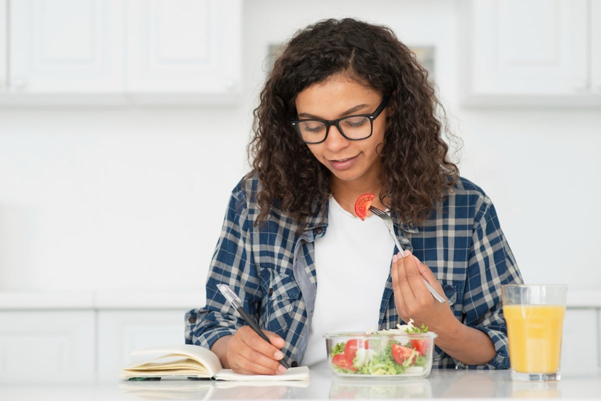 Alimentação Equilibrada Durante os Estudos - Preparação para o Enem 2025 Estudante de óculos se alimentando com salada saudável enquanto estuda com caderno aberto na cozinha durante preparação para Enem