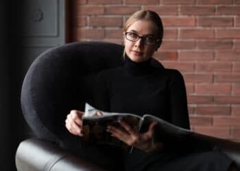 Profissional de óculos lendo revista especializada sobre psicopatia corporativa sentada em poltrona moderna