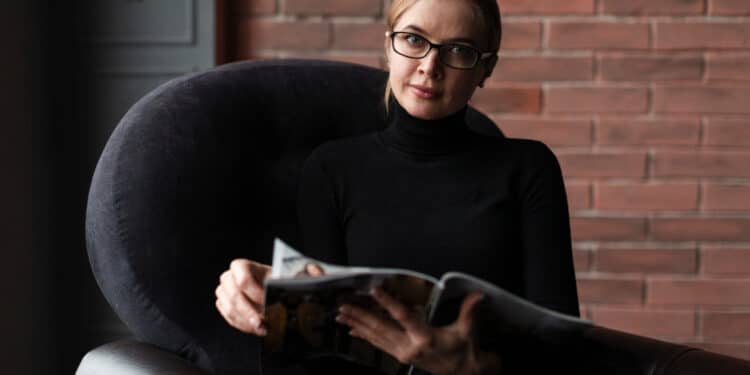 Profissional de óculos lendo revista especializada sobre psicopatia corporativa sentada em poltrona moderna