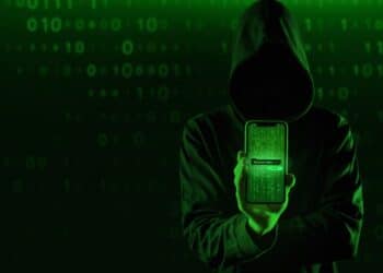 Figura encapuzada segurando smartphone com código verde matrix na tela representando cibercriminoso aplicando golpe