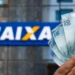 Mão segurando cédulas de 100 reais na frente da fachada com logo da Caixa Econômica