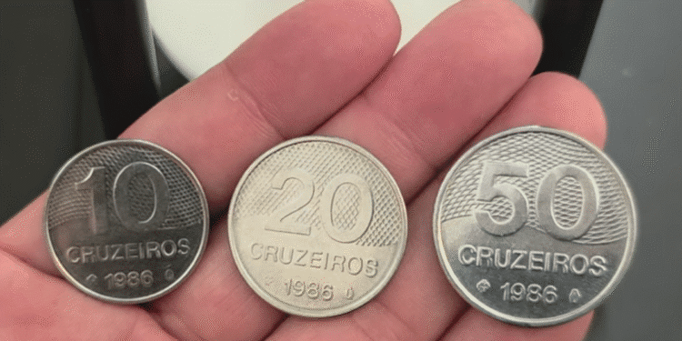 Três moedas antigas de 10, 20 e 50 Cruzeiros do ano de 1986, sendo seguradas na palma da mão, em estado de Flor de Cunho. Imagem ilustra a raridade das moedas de baixa tiragem procuradas por colecionadores de numismática.