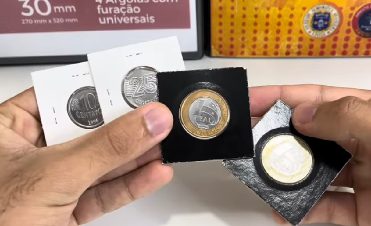 Mãos de um colecionador exibindo quatro moedas raras do Plano Real (incluindo R$ 1 e R$ 0,25) em protetores numismáticos, destacando o estado de conservação.