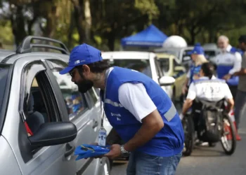 Homem de colete azul entregando materiais para motorista em carro cinza durante ação comunitária ao ar livre