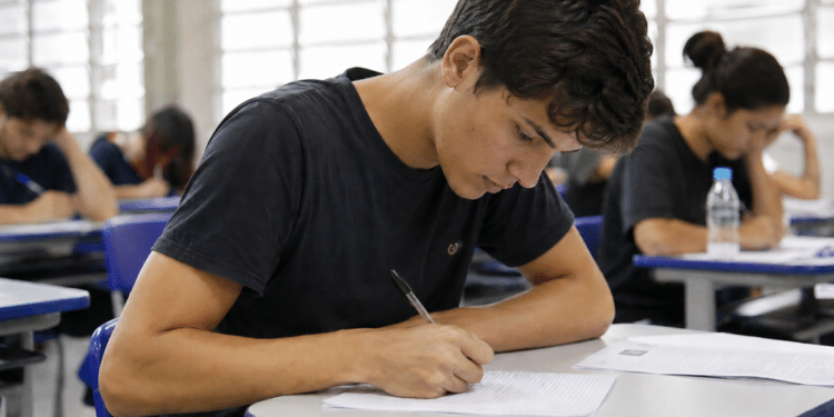 Rapaz escrevendo concentrado em prova, rodeado de outros jovens em sala de aula, mesas azuis.