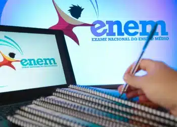 Logo do Enem em computador e tela de fundo, com a mão de um estudante com caneta sobre cadernos. Imagem relacionada à preparação e estudo para a prova de Redação do Exame Nacional do Ensino Médio.